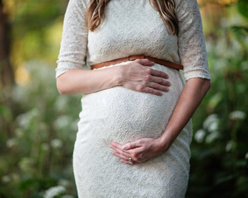 A pregnant woman in a lace dress gently cradling her belly in an outdoor setting.